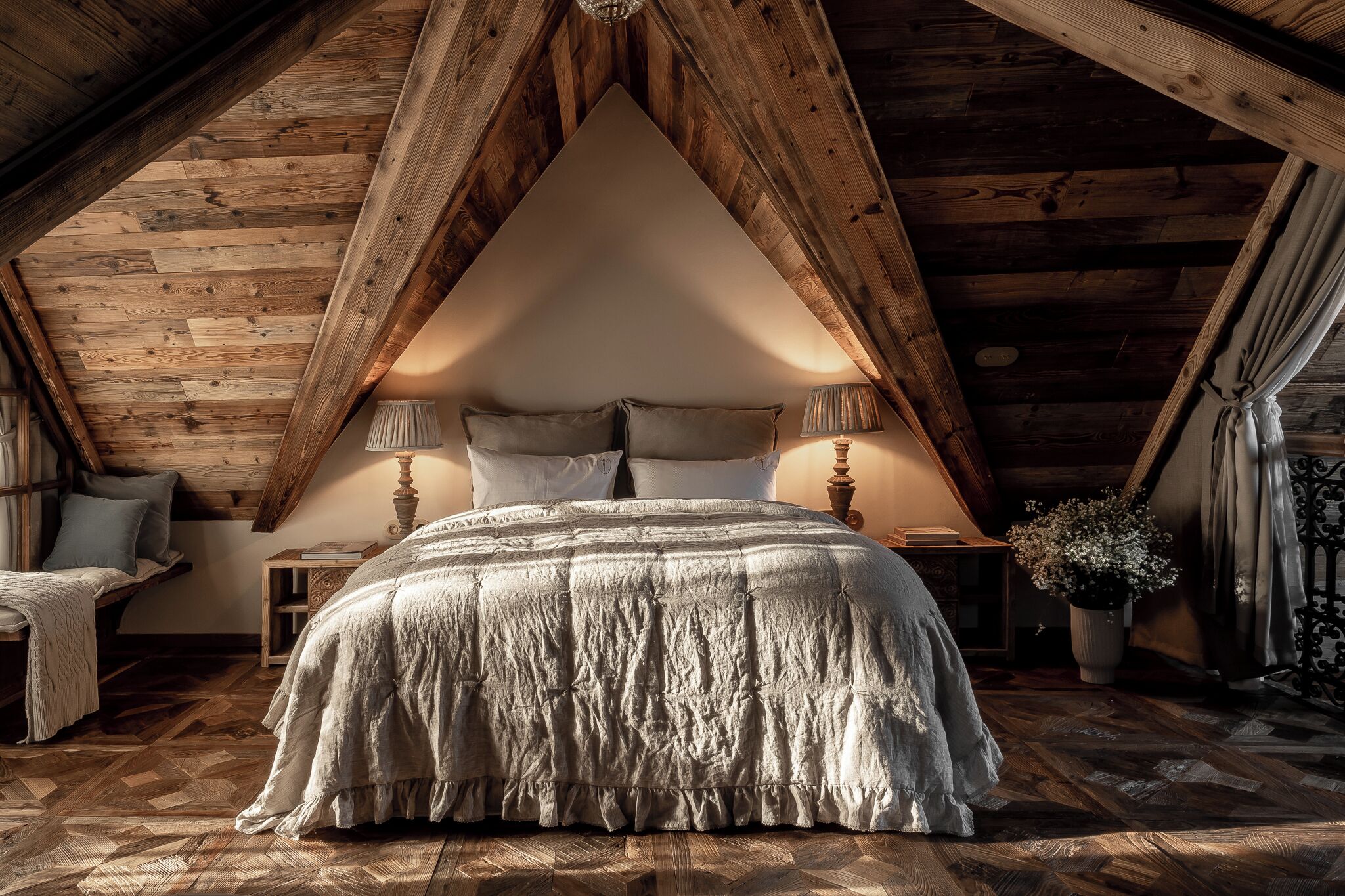 Un dormitorio con techo abovedado, vigas de madera, una cama grande con una manta texturizada, lámparas de noche y una planta en maceta.