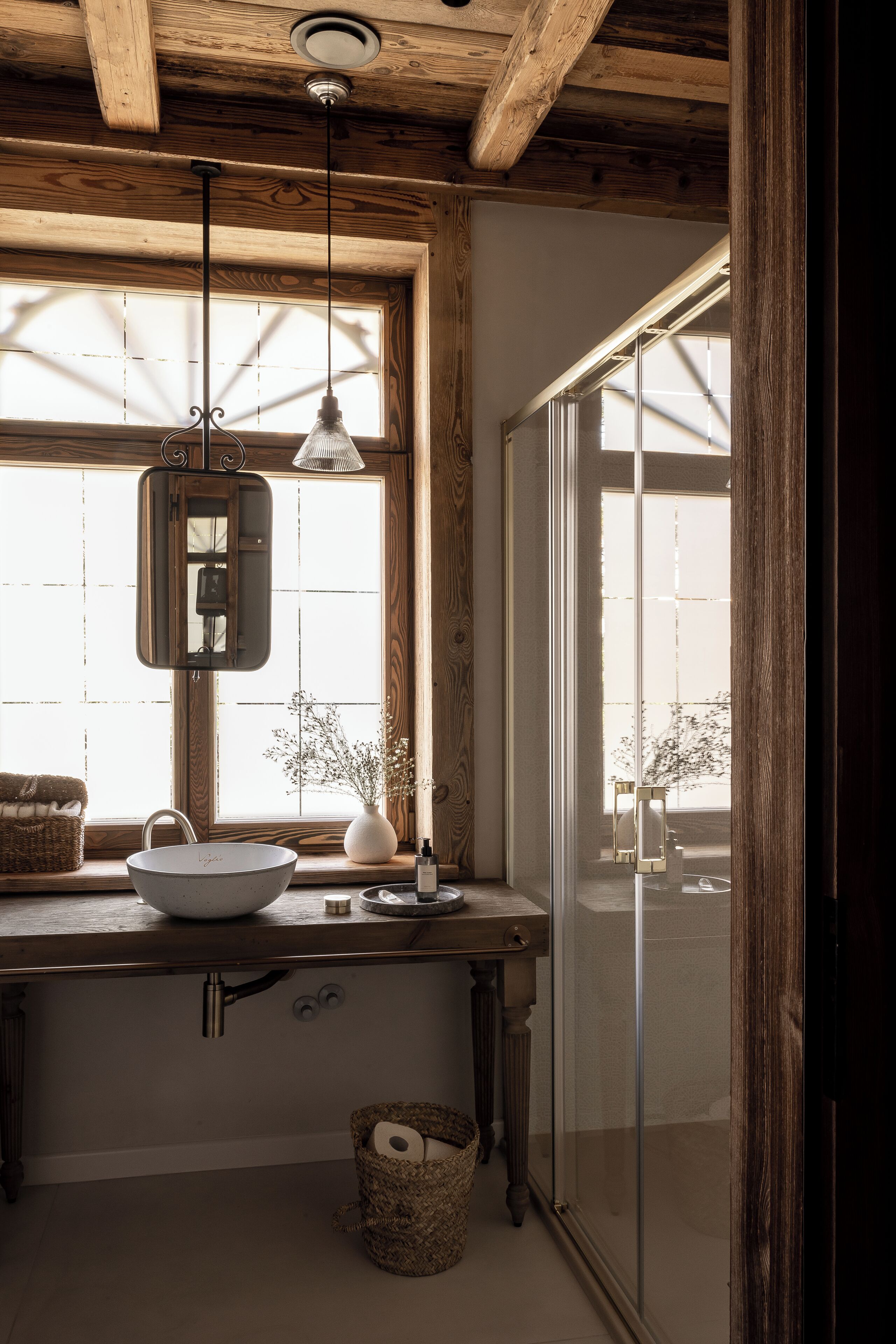 A rustic bathroom with a wooden ceiling, a large window with a view, a sink with a mirror, and a hanging light fixture.