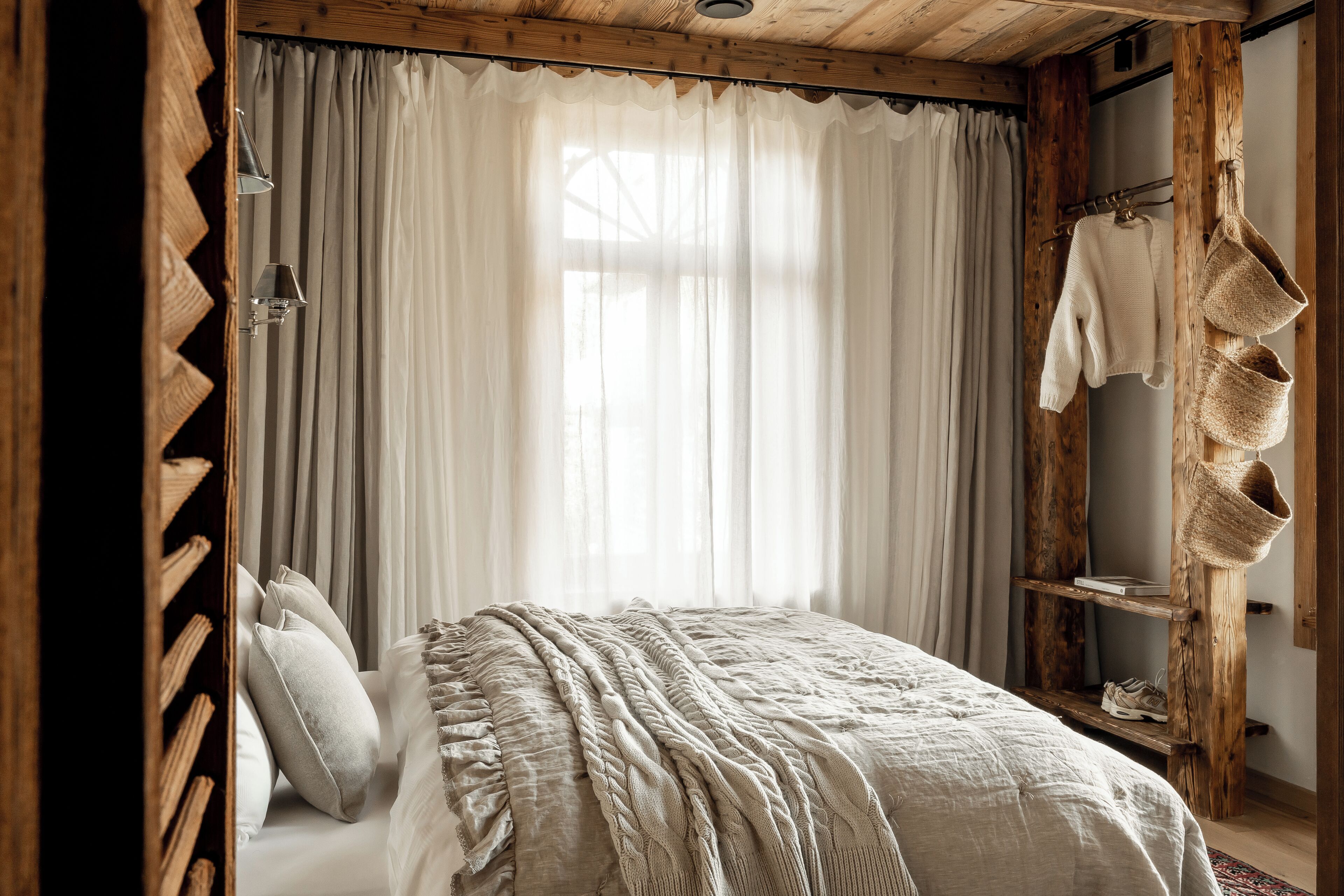 Un dormitorio con cama, cortinas, un estante de madera con zapatos y una cesta colgante.