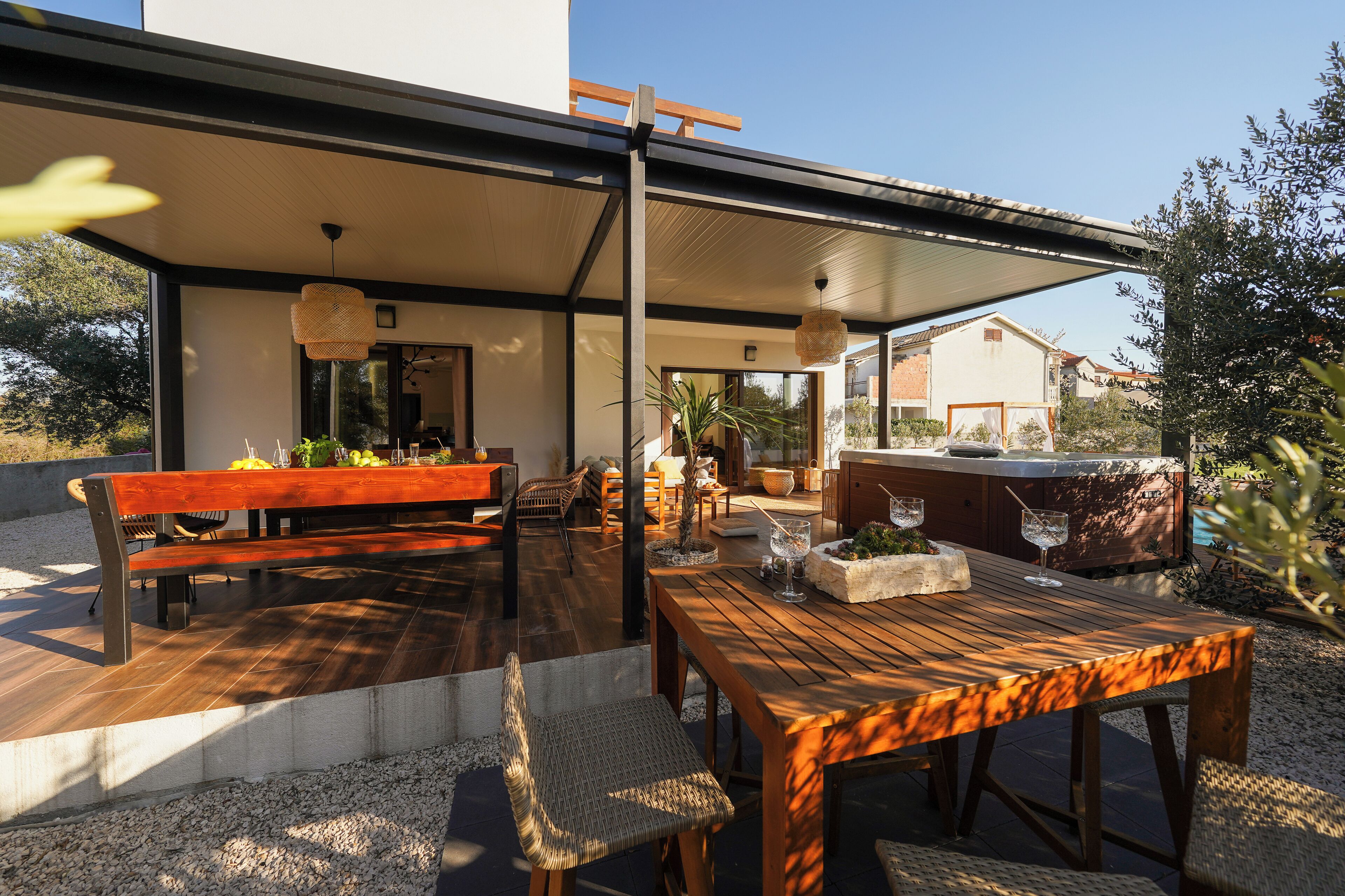 Un patio exterior con una mesa y bancos de madera, rodeado de plantas y una zona de barbacoa.