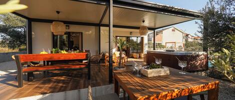 Un patio exterior con una mesa y bancos de madera, rodeado de plantas y una zona de barbacoa.