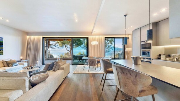 A modern living room with a sofa, coffee table, and chairs, a kitchen area with bar stools, and large windows offering a view of the sea. - Luxury Lakefront Loft in Montreux Swiss Reviera (Territet)