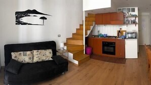 A modern apartment with a black sofa, wooden staircase, kitchen, dining area, and a wall with a black and white mural.
