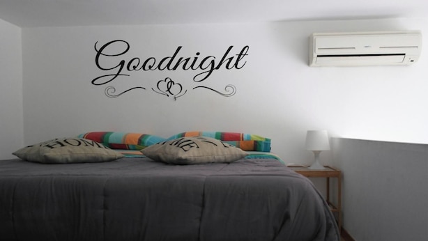 A bedroom with a bed, pillows, a bedside table, a lamp, and a wall-mounted air conditioner.