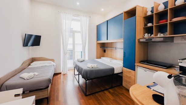 A compact hotel room with two single beds, a desk with a computer, a small kitchenette, and a TV mounted on the wall. - Ale & Tizy House 2 Studio (Napoli)