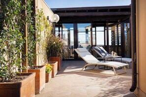 A sunlit outdoor patio with lounge chairs, potted plants, and a view of the city.