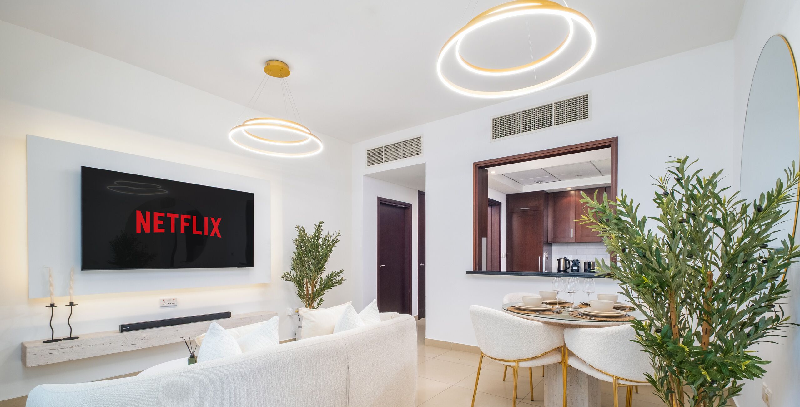 A modern living room with a flat-screen TV, a sofa, a dining area with a table set for four, and a large mirror.