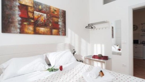 A hotel room with a bed, a painting on the wall, a small table with a red object, and a mirror. - Jade 103 - Front of SMN Station - 2BR (Firenze)