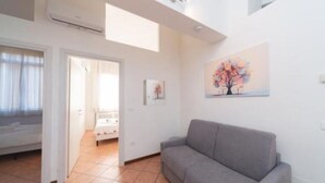 A spacious living area with a grey sofa, a painting on the wall, and a door leading to another room. - Jade 103 - Front of SMN Station - 2BR (Firenze)