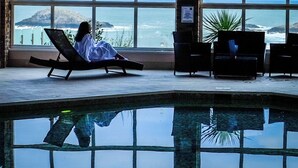 Una persona descansando en una silla de descanso junto a un ventanal grande con vista a una piscina y al océano.