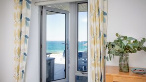 Un balcón con vistas al mar, un mueble de madera con un florero de follaje, y una cortina estampada.