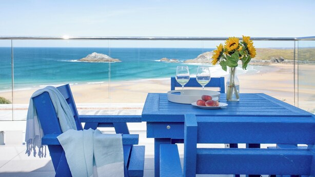 Una mesa azul con un jarrón de girasoles, dos copas de vino y un plato de fruta en un balcón con vistas al océano.