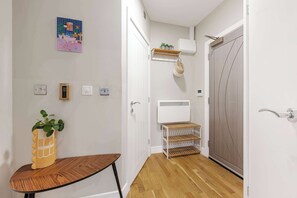 A hallway with a wooden floor, a white door, a wall-mounted shelf with plants, a wall-mounted air conditioner, and a small table with a plant.