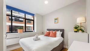 A bedroom with a large bed, a window with a view of a building, a bedside table with a lamp, and a potted plant. - 1 Bed Northern Quarter Loft | City Centre | Air Conditioned (Manchester)