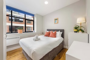 A bedroom with a large bed, a window with a view of a building, a bedside table with a lamp, and a potted plant.