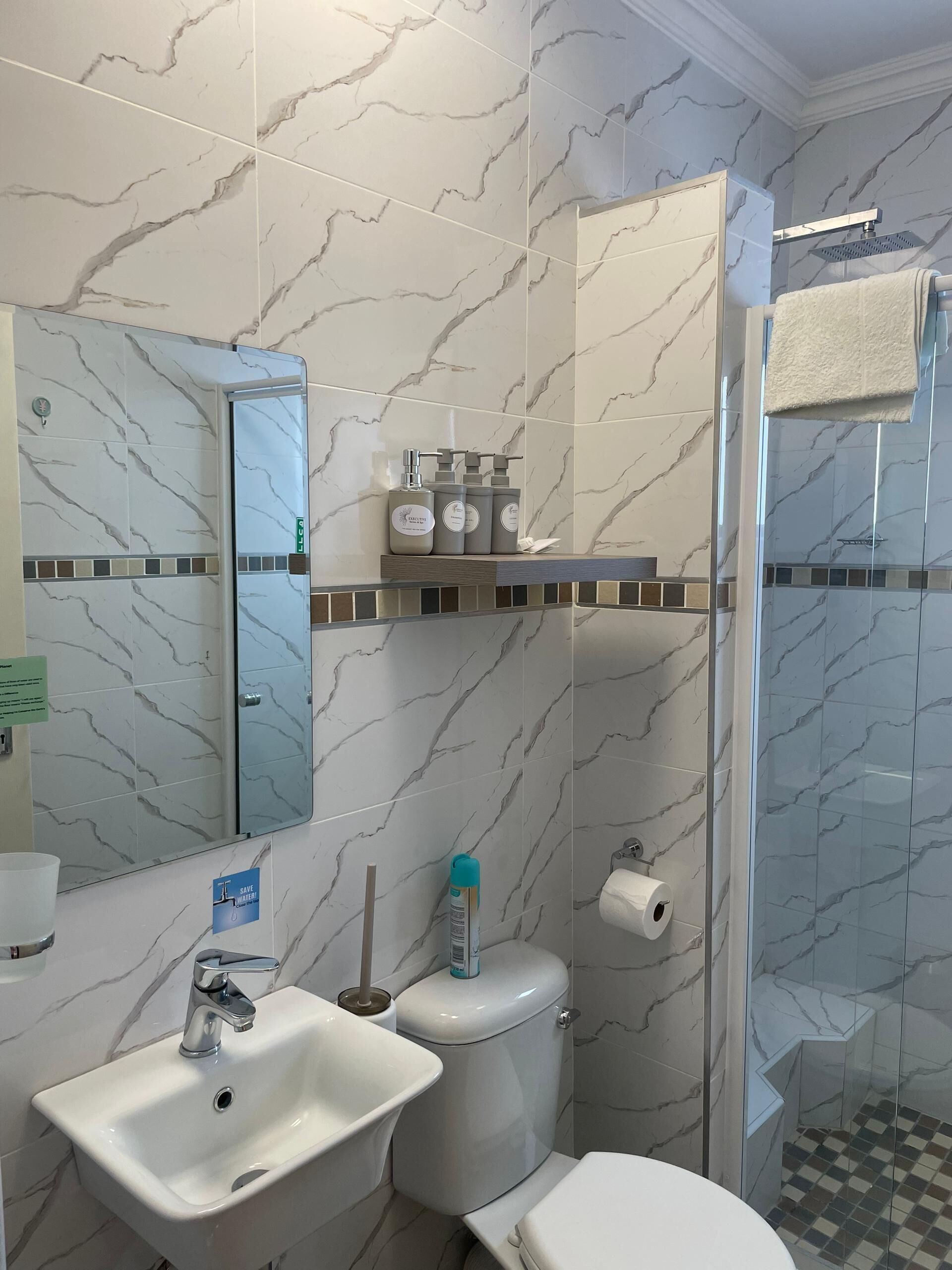 A bathroom with marble walls, a sink, a toilet, a shower, and toiletries on a shelf.