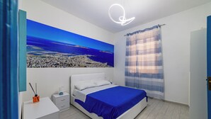 A bedroom with a bed, a nightstand, a desk, a large window with curtains, and a cityscape painting on the wall.