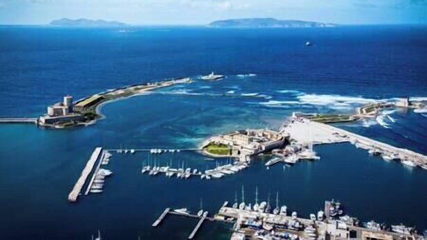 An aerial view of a marina with boats docked, a pier extending into the water, and a small island with buildings.