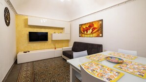 A modern living room with a flat-screen TV, a sofa, a dining table with placemats, and a patterned floor.