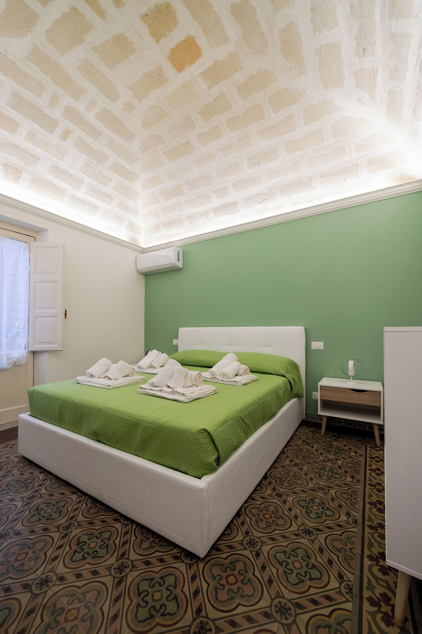 A room with a green wall, a white bed with green bedding, a patterned floor, and a white ceiling with a brick-like texture.