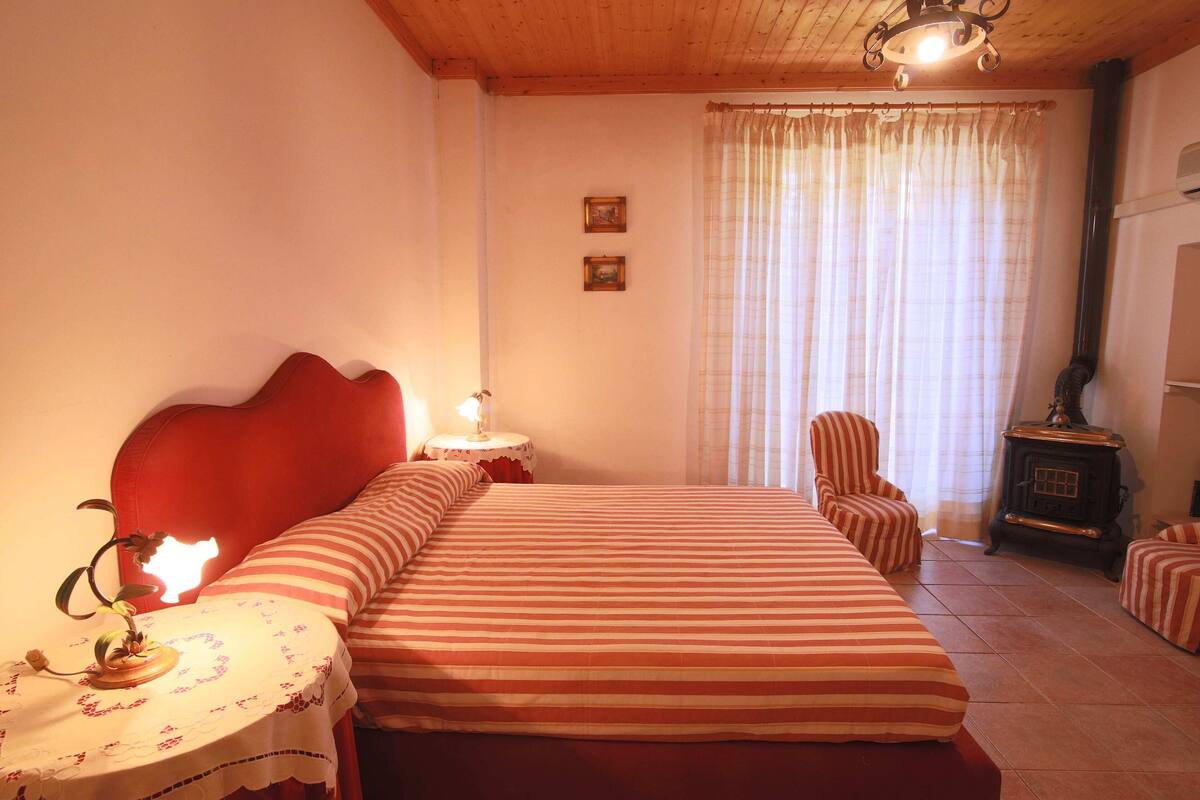 Une pièce confortable avec un grand lit, une petite table avec une lampe, une chaise et un poêle à bois.