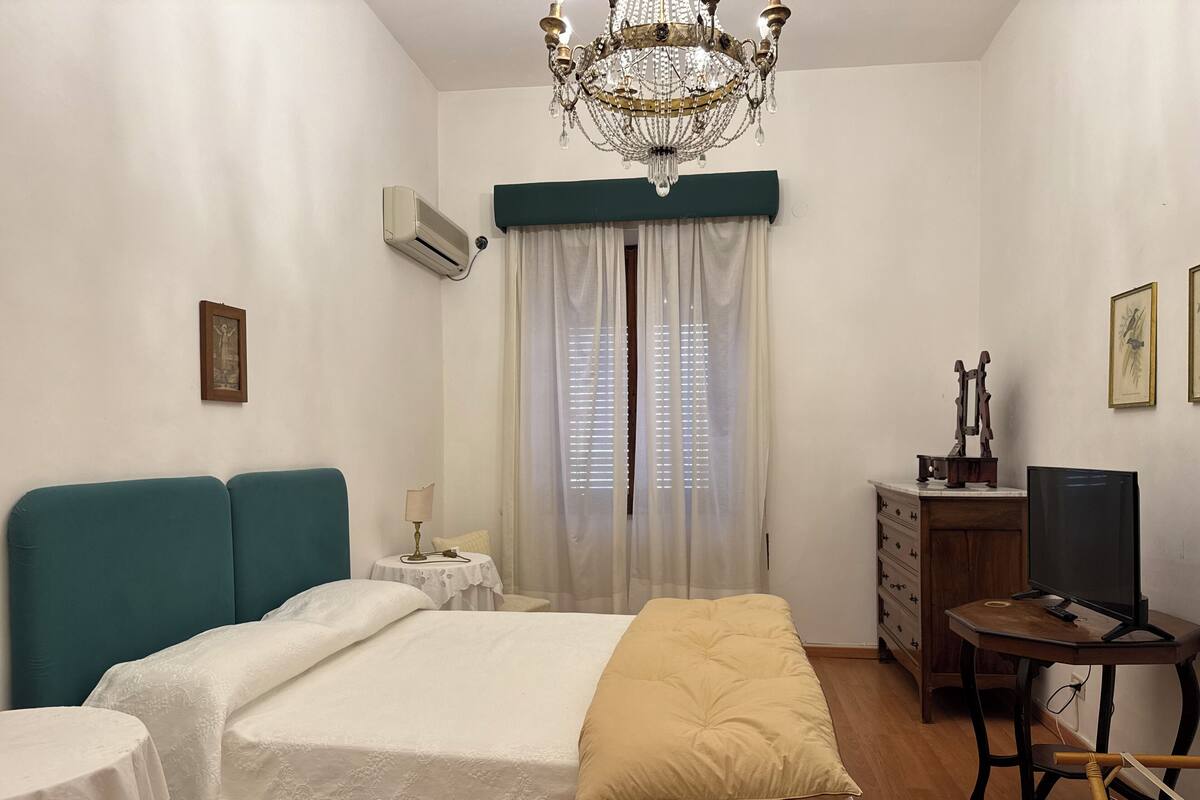 Une chambre à coucher avec un lit, un bureau, une chaise, un lustre et une fenêtre avec des rideaux.