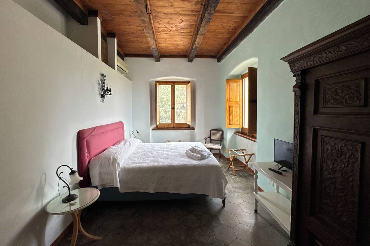 Une chambre à coucher comprenant un lit, une chaise, une petite table, un téléviseur et une fenêtre avec des volets en bois.