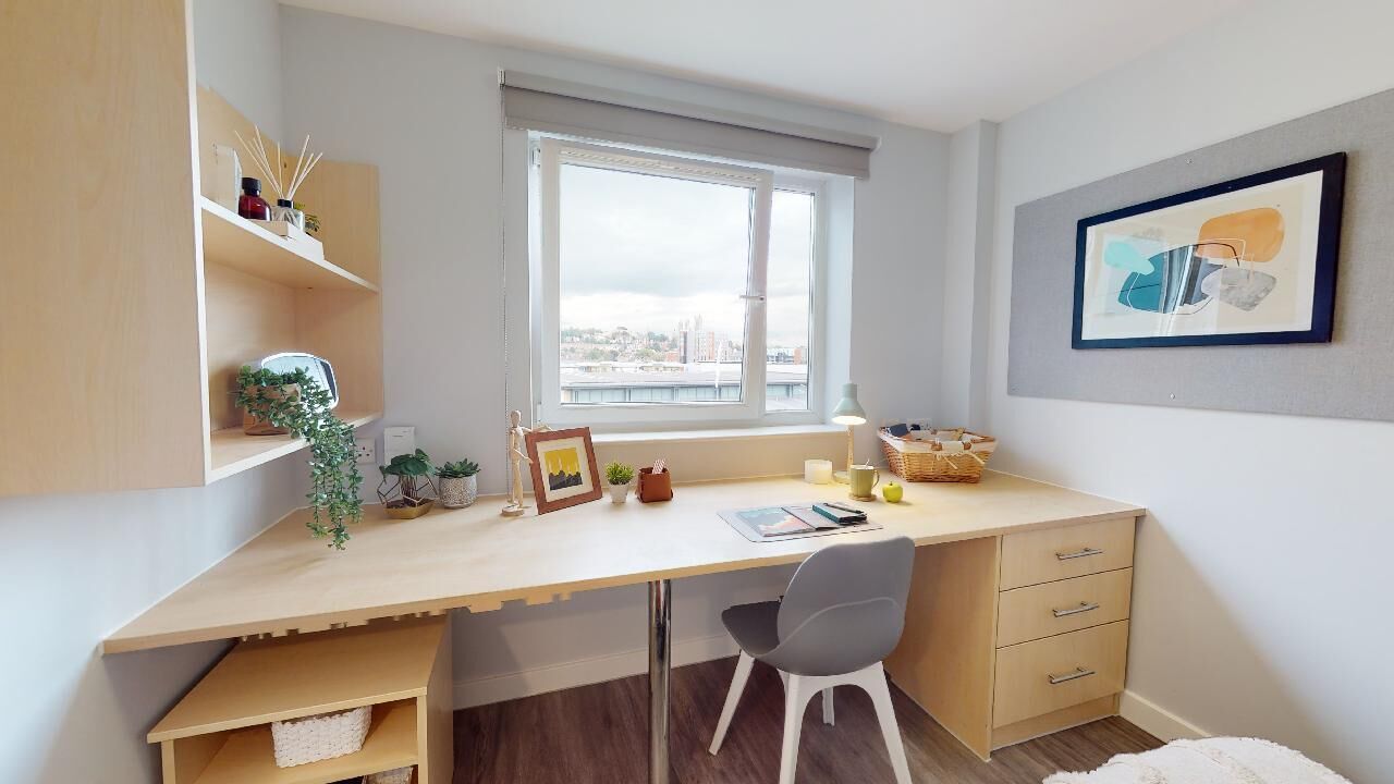 Un dormitorio moderno con un escritorio grande, una silla, una ventana con vista a la ciudad, una mesita de noche y una cama.