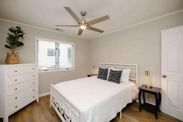 Une chambre à coucher comprenant un lit, un ventilateur de plafond, une fenêtre avec des stores, une commode et une table de chevet avec une lampe.