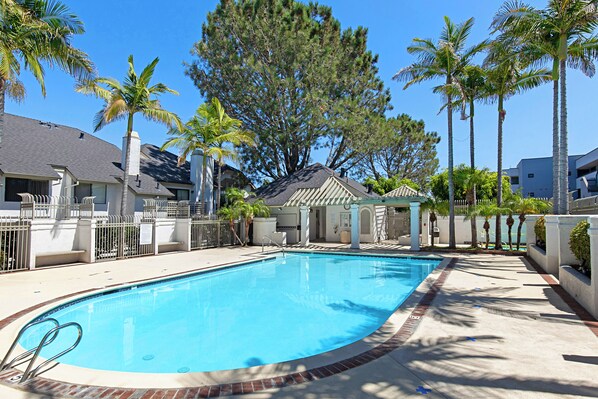 A swimming pool with a brick border, surrounded by palm trees and a covered seating area.