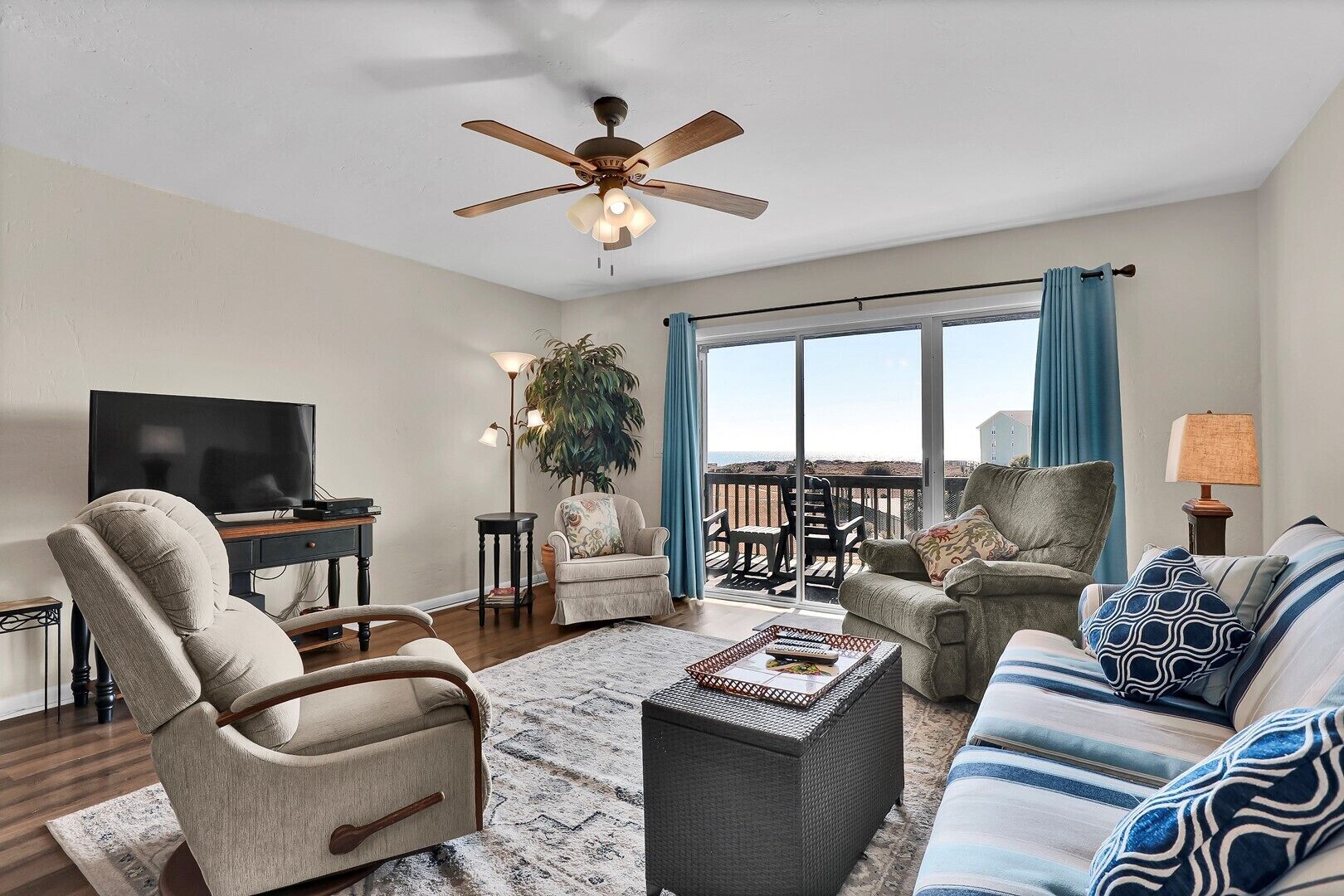 A spacious living room with a large TV, comfortable seating, a ceiling fan, and a balcony with a view.