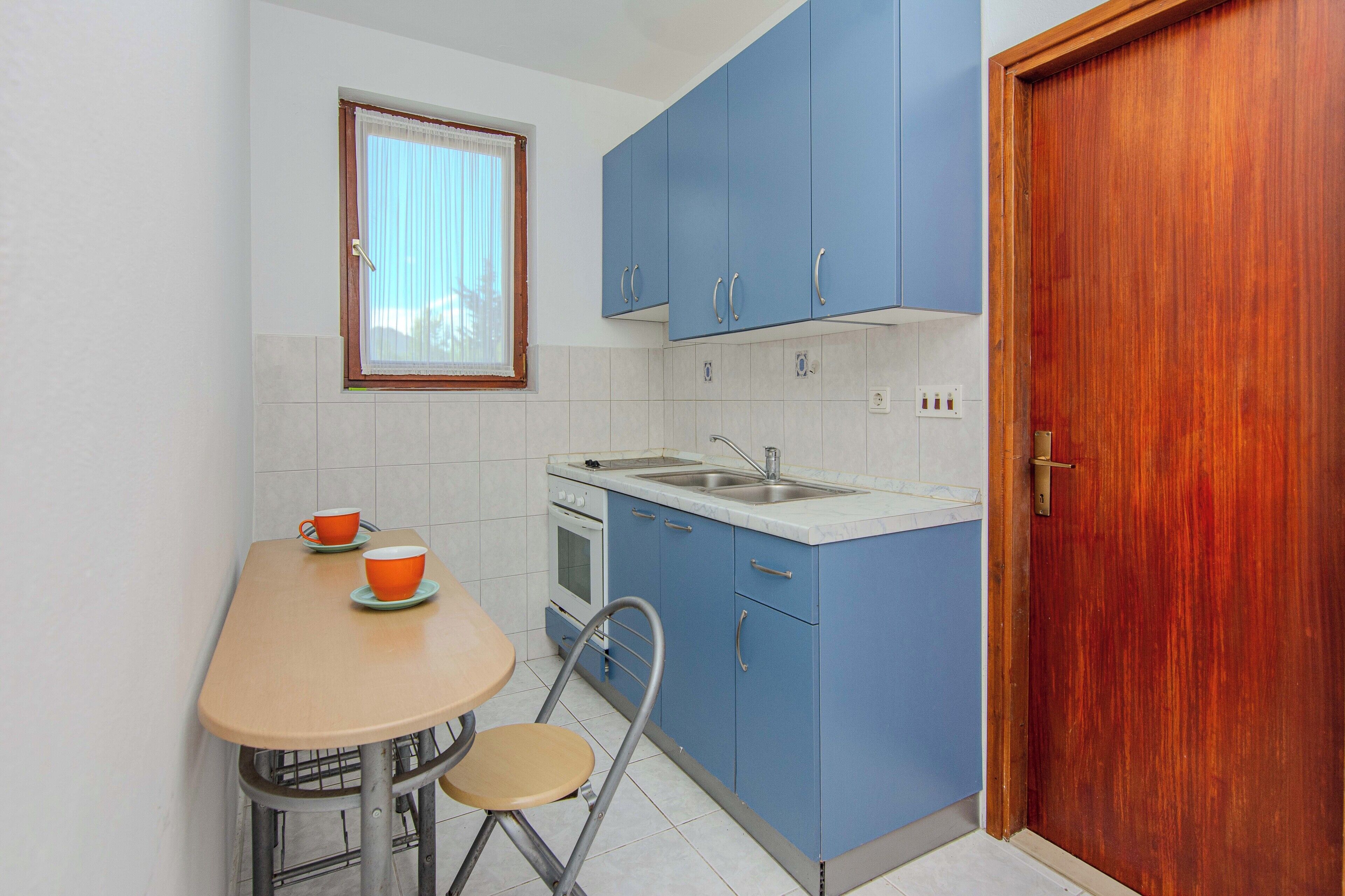 Una pequeña cocina con armarios azules, una puerta de madera, una ventana con cortinas, un lavamanos, un horno y una mesa con dos tazas naranjas.