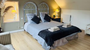 A bedroom with a large bed, two bedside tables, a radiator, and a window with a lamp. - Le "33" Gîte de Charme coeur Cotentin (Valognes)