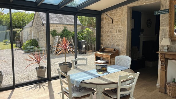A room with a large glass door leading to a garden, a wooden table with chairs, a stone wall, and a small desk with a lamp. - Le "33" Gîte de Charme coeur Cotentin (Valognes)