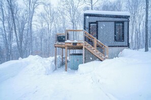 Une cabane enneigée, dotée d’une terrasse en bois et d’une fenêtre, entourée d’arbres.