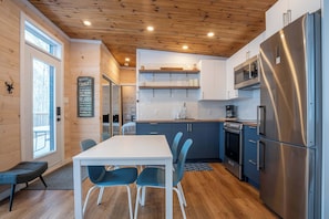 A modern kitchen with wooden ceiling, stainless steel appliances, and a dining area with a white table and blue chairs.