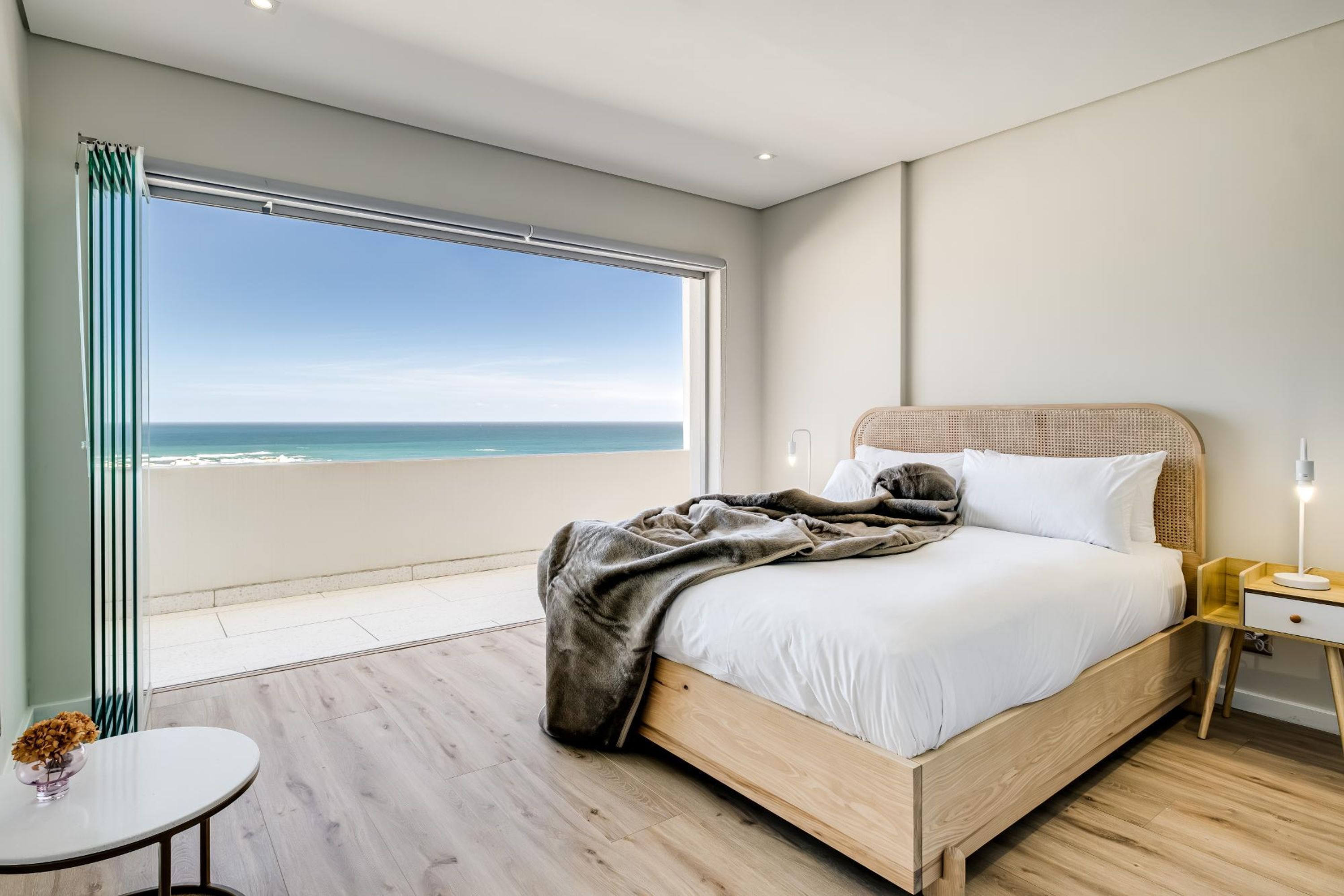 Une chambre avec une grande fenêtre donnant sur la mer, un lit avec une couverture, une table de chevet avec une lampe et une petite table avec un vase.