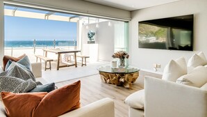 A modern living room with a large TV, comfortable seating, a glass coffee table, and a view of the ocean. - Clifton 2bed Penthouse Sea Views Dunmore Horizons (Cape Town)