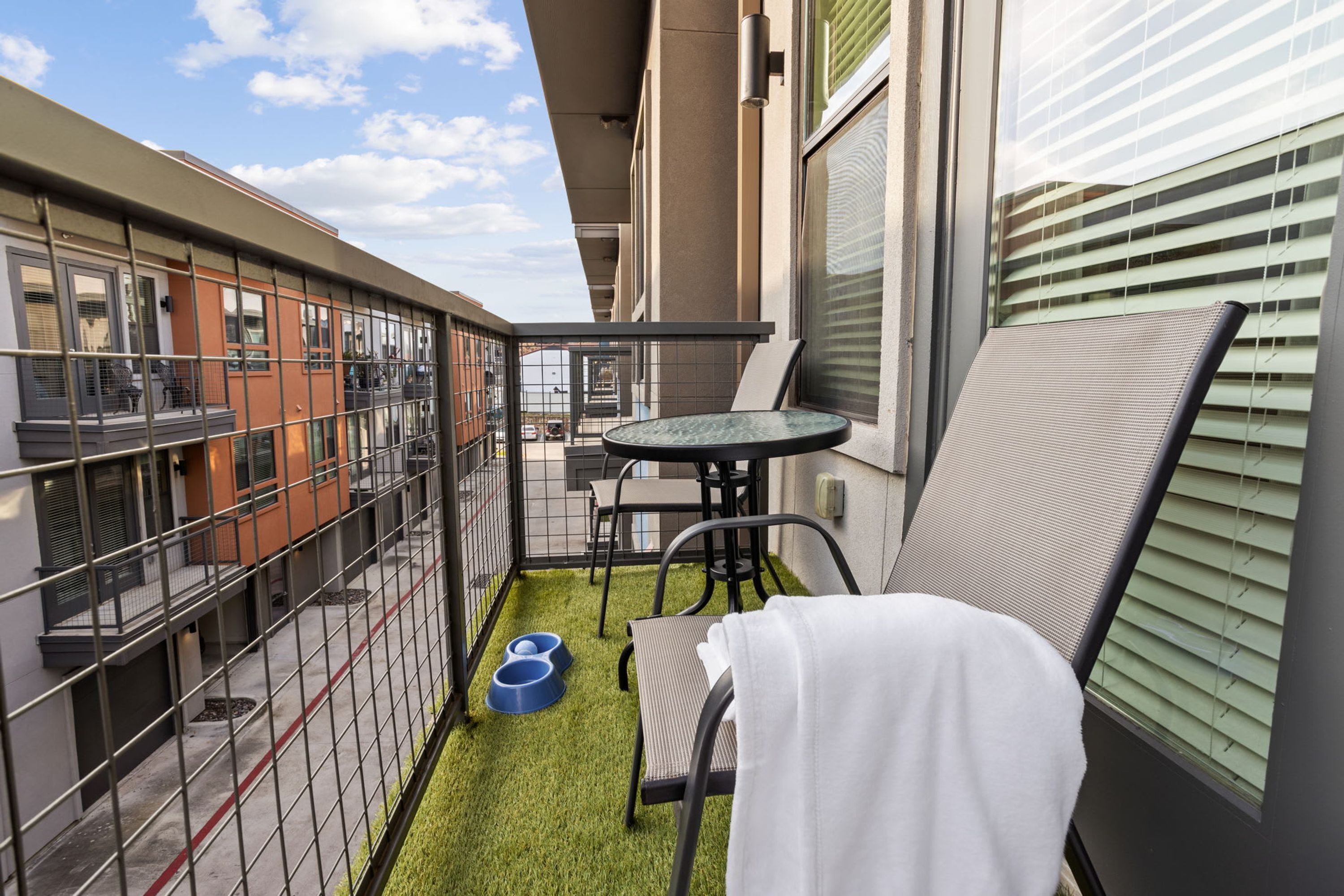 A balcony with a small table and chairs, a pet feeding station, and a view of neighboring buildings.