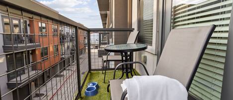 A balcony with a small table and chairs, a pet feeding station, and a view of neighboring buildings.