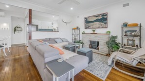 Ein modernes Wohnzimmer mit grauem Sofa, Holzboden, Deckenventilator, Kamin und einem Essbereich mit weißem Tisch und Stühlen.