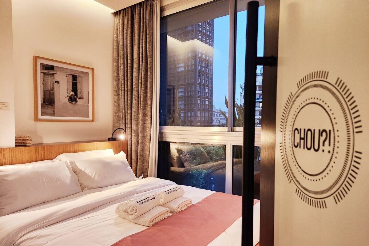 Una habitación de hotel con cama, ventana con vista a edificios, un cuadro en la pared y un adorno mural con la palabra "CHOU!".