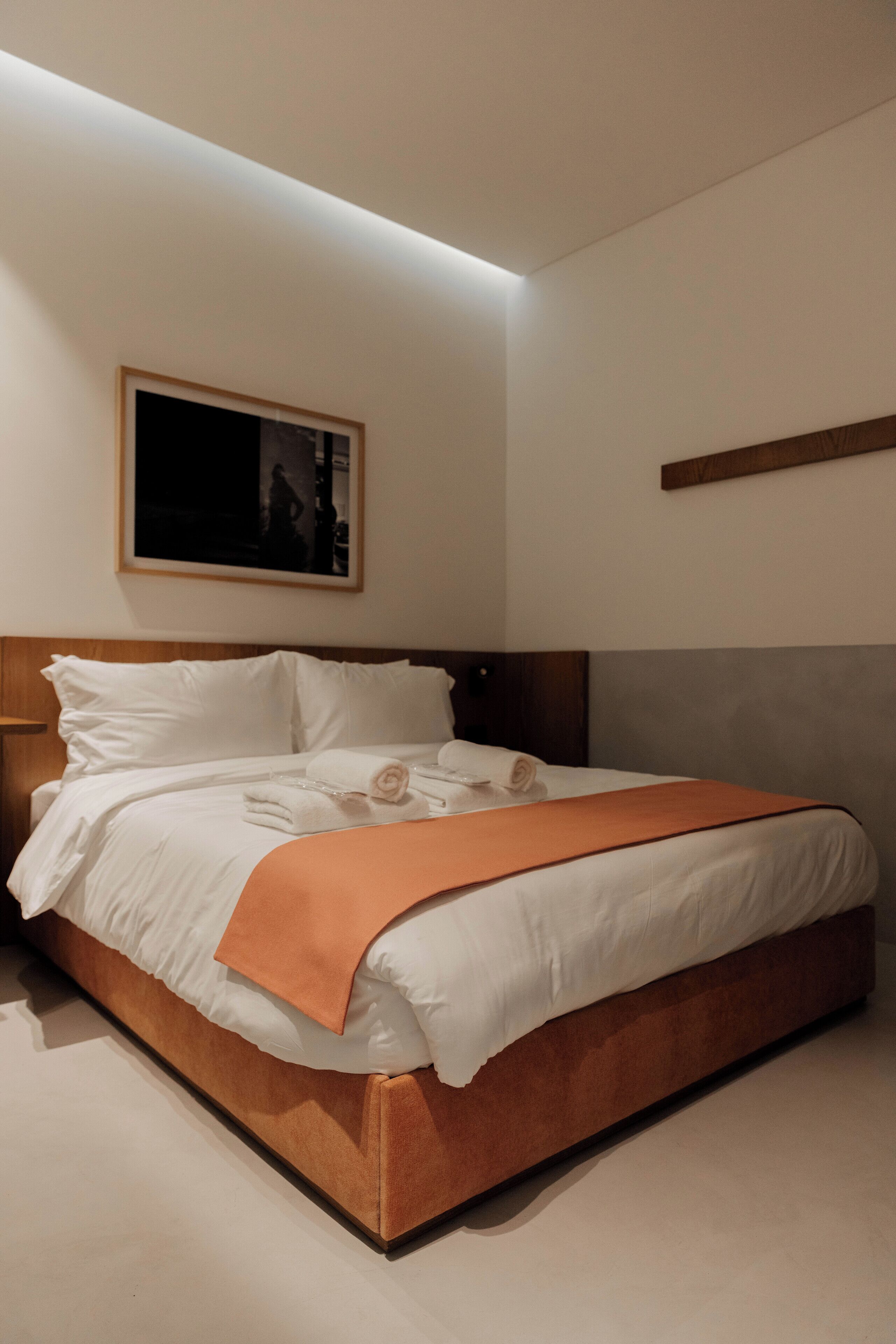 A neatly made bed with white linens and an orange throw blanket, a wooden headboard, and a framed picture on the wall.