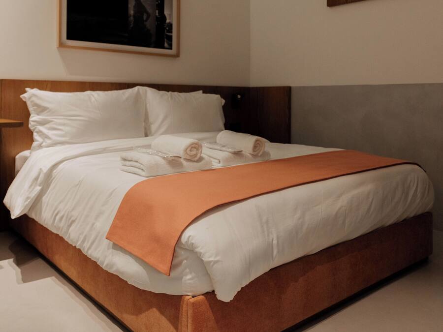A neatly made bed with white linens and an orange throw blanket, a wooden headboard, and a framed picture on the wall.