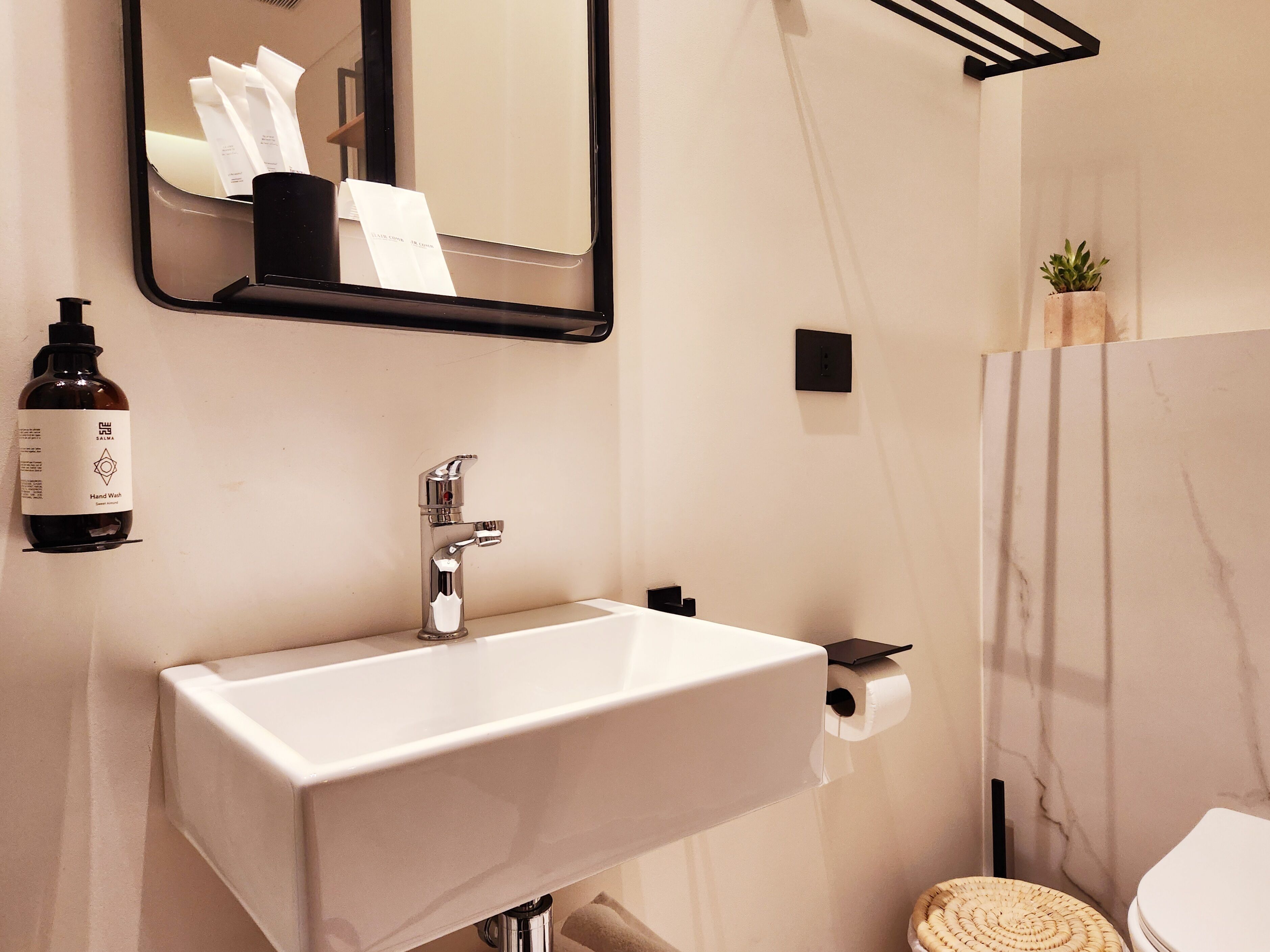 A bathroom with a white sink, a mirror, a bottle of hand wash soap, a towel, and a toilet.
