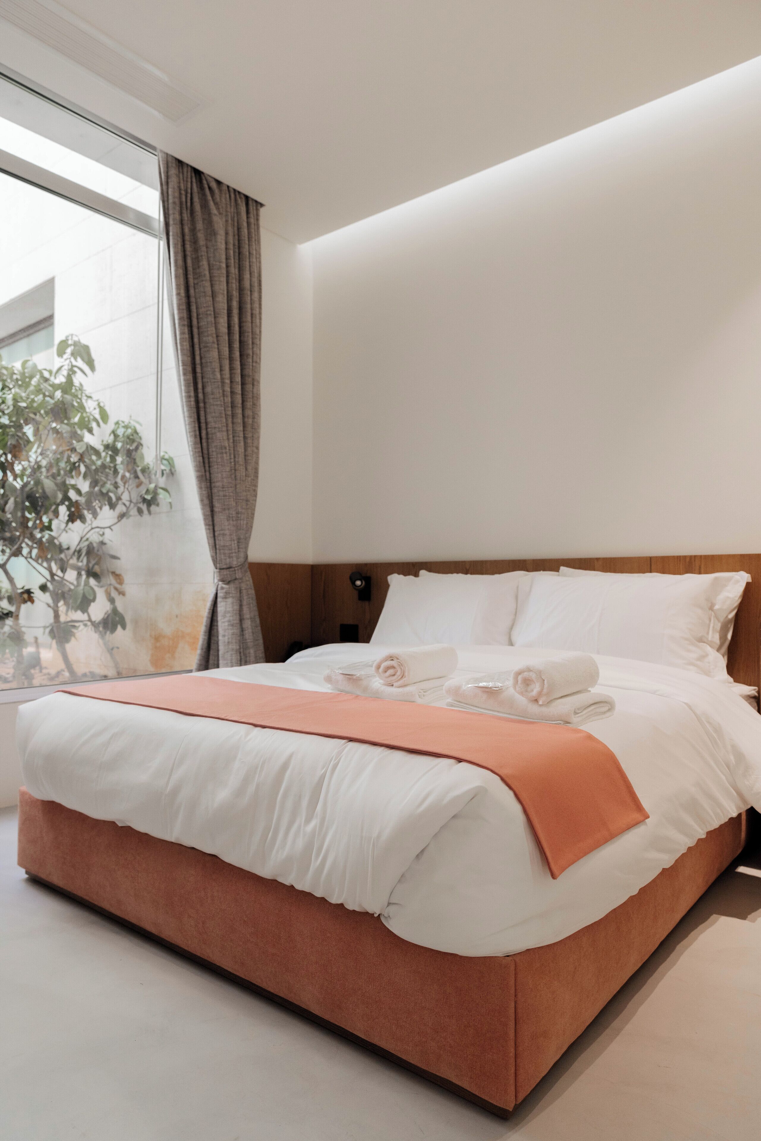 Quarto de hotel moderno com uma cama grande, cabeceira de madeira e vista para uma árvore pela janela.