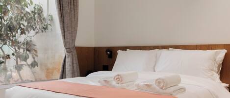 Quarto de hotel moderno com uma cama grande, cabeceira de madeira e vista para uma árvore pela janela.