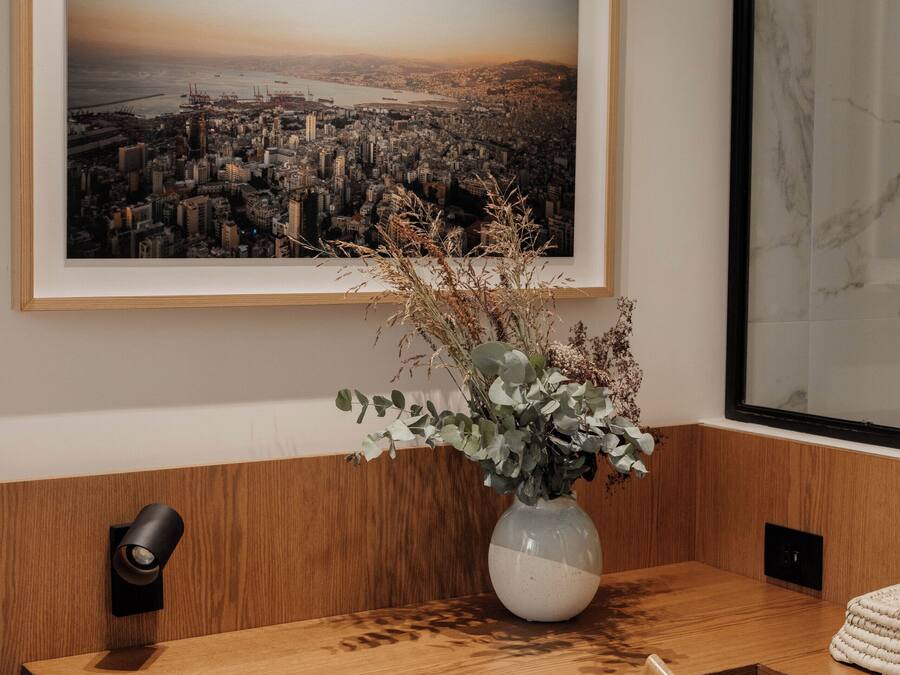 A wooden desk with a vase of dried plants, a framed cityscape picture, and a marble wall.