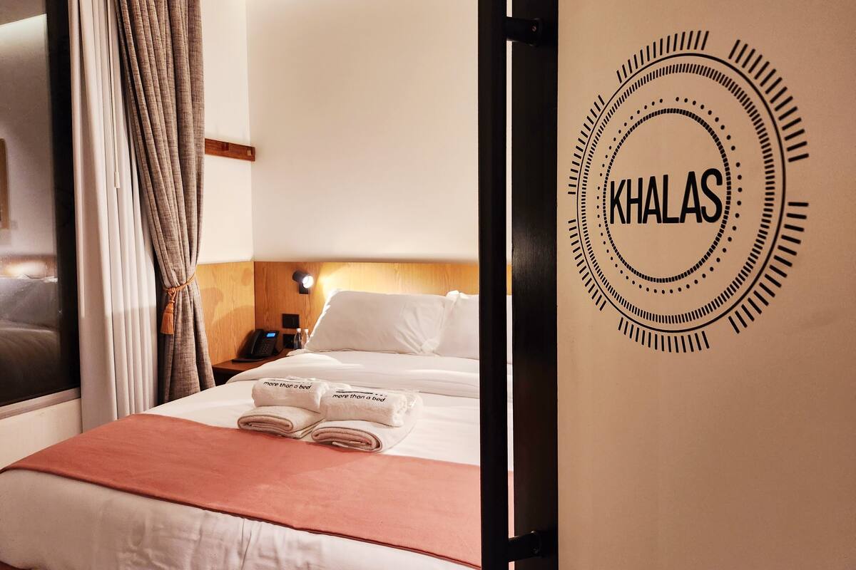 Una habitación de hotel con una cama bien hecha, una mesita de noche con lámpara y una pared con el nombre del hotel 'KHALAS'.