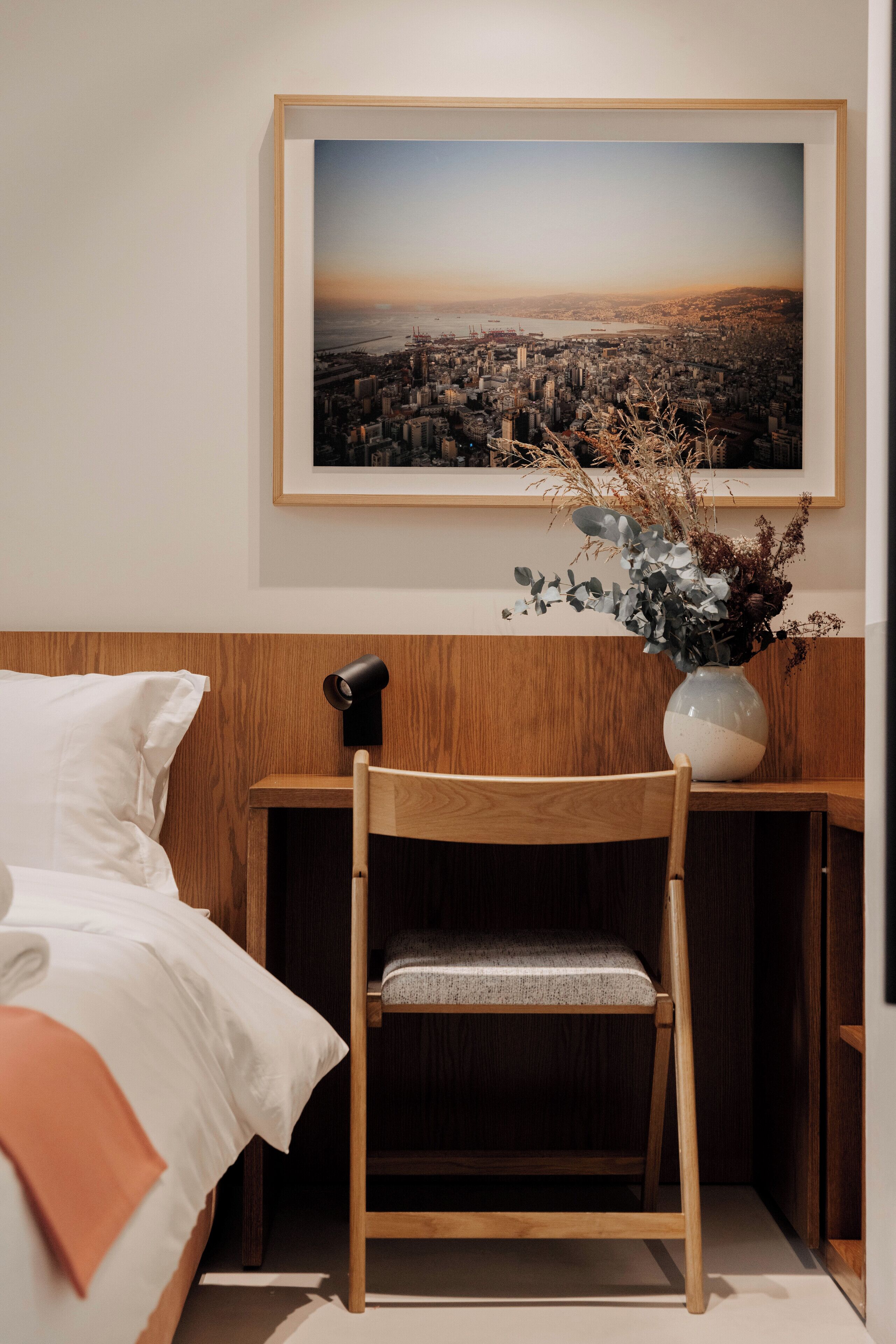 Une chambre d’hôtel avec un lit, une chaise en bois, un bureau avec une lampe, un vase contenant des plantes séchées et, au mur, un tableau encadré représentant un paysage urbain.